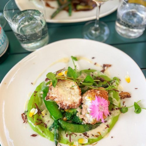 Un plat de légumes et cromesquis avec sauce, décoré de fleurs comestibles et accompagné d'un verre de vin blanc.