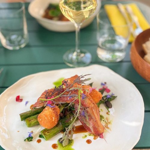 Un plat de légumes et viande avec sauce, décoré de fleurs comestibles et accompagné d'un verre de vin blanc.